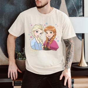 Puede incluir: Camiseta color crema con una ilustraci&oacute;n de dibujos animados de dos personajes femeninos con cabello rubio y casta&ntilde;o rojizo, rodeados de estrellas. Un personaje viste un atuendo azul, el otro una capa rosa. El texto "Disney" es visible.