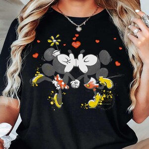 May include: Black t-shirt featuring a graphic of two kissing cartoon mice with yellow shoes and red heart accents. The design includes a flower and yellow paint splatters. The shirt is a casual wear item.