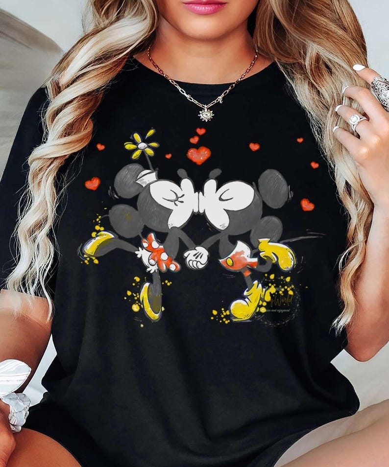 May include: Black t-shirt featuring a graphic of two kissing cartoon mice with yellow shoes and red heart accents. The design includes a flower and yellow paint splatters. The shirt is a casual wear item.