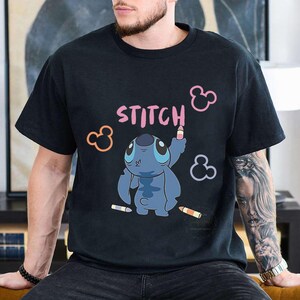 May include: Black t-shirt featuring a cartoon Stitch character drawing the word "STITCH" in pink. The design includes pink, orange, and purple Mickey Mouse ear shapes and colored pencils. The shirt is a casual, unisex style.