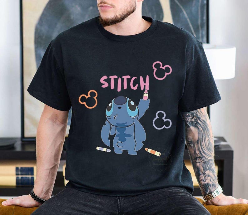 May include: Black t-shirt featuring a cartoon Stitch character drawing the word "STITCH" in pink. The design includes pink, orange, and purple Mickey Mouse ear shapes and colored pencils. The shirt is a casual, unisex style.