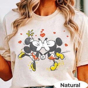 May include: A beige t-shirt featuring a graphic of Mickey and Minnie Mouse kissing, surrounded by red hearts and a flower. Mickey Mouse is wearing yellow shoes and red shorts, while Minnie Mouse wears a polka-dot skirt. The word "Natural" is printed at the bottom.