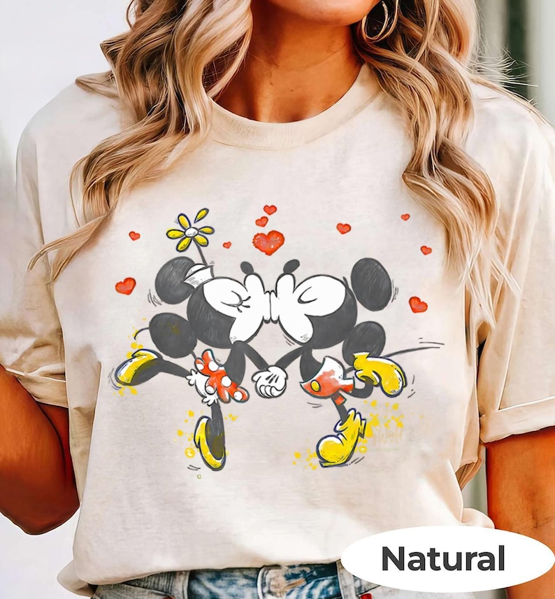 May include: A beige t-shirt featuring a graphic of Mickey and Minnie Mouse kissing, surrounded by red hearts and a flower. Mickey Mouse is wearing yellow shoes and red shorts, while Minnie Mouse wears a polka-dot skirt. The word "Natural" is printed at the bottom.