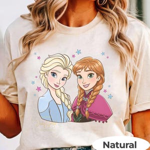 Puede incluir: Camiseta beige con una ilustraci&oacute;n de dibujos animados de dos personajes femeninos con cabello rubio y casta&ntilde;o rojizo, rodeados de estrellas. Los personajes visten atuendos azules y rosas. El texto "Disney World" es visible. La camiseta tiene una etiqueta "Natural".