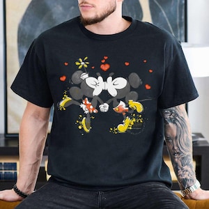 May include: Black t-shirt featuring a graphic of Mickey and Minnie Mouse kissing, surrounded by red hearts and a yellow flower. The cartoon characters are in black, white, red, and yellow. The shirt is short-sleeved.