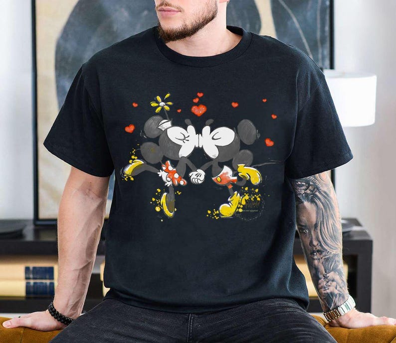 May include: Black t-shirt featuring a graphic of Mickey and Minnie Mouse kissing, surrounded by red hearts and a yellow flower. The cartoon characters are in black, white, red, and yellow. The shirt is short-sleeved.