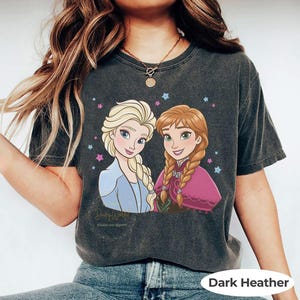 Puede incluir: Camiseta gris oscuro con un gr&aacute;fico de Elsa y Anna de Frozen, con estrellas. La camiseta tiene cuello redondo y mangas cortas. El texto "DisneyWorld" est&aacute; impreso en la camiseta.