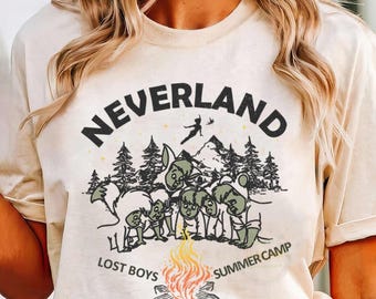 May include: A beige t-shirt with the word "NEVERLAND" in black block letters. Below the text is a graphic of the Lost Boys around a campfire, with the words "LOST BOYS SUMMER CAMP". The shirt is labeled "Natural".