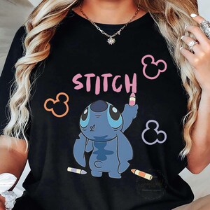 May include: Black t-shirt featuring the word "STITCH" in pink, with a blue cartoon character holding a crayon. Mickey Mouse silhouettes in pink, orange, and purple surround the design. Two crayons are at the bottom.