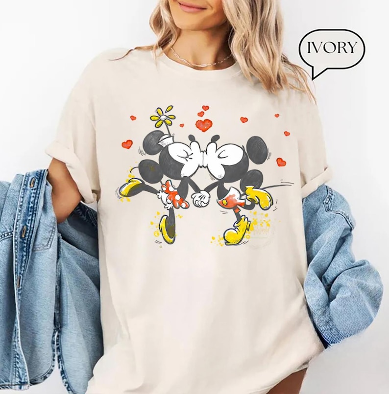 May include: Ivory-colored t-shirt featuring a cartoon illustration of Mickey and Minnie Mouse kissing, surrounded by red hearts and a flower. The text "IVORY" is in a speech bubble. The shirt has short sleeves.