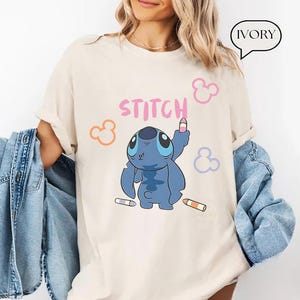 May include: Ivory-colored t-shirt featuring a blue cartoon character, holding a pink crayon. The word "STITCH" is written in pink above the character. Orange, pink, and purple Mickey Mouse-style shapes are scattered around the design.