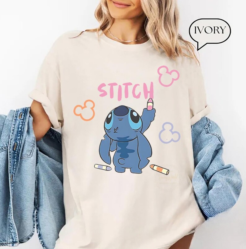 May include: Ivory-colored t-shirt featuring a blue cartoon character, holding a pink crayon. The word "STITCH" is written in pink above the character. Orange, pink, and purple Mickey Mouse-style shapes are scattered around the design.