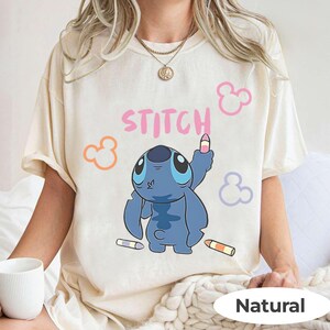 May include: Cream-colored t-shirt featuring a blue cartoon character holding a crayon, with the word "STITCH" in pink. The design includes colorful crayon accents and Mickey Mouse-like shapes. The shirt is labeled "Natural".