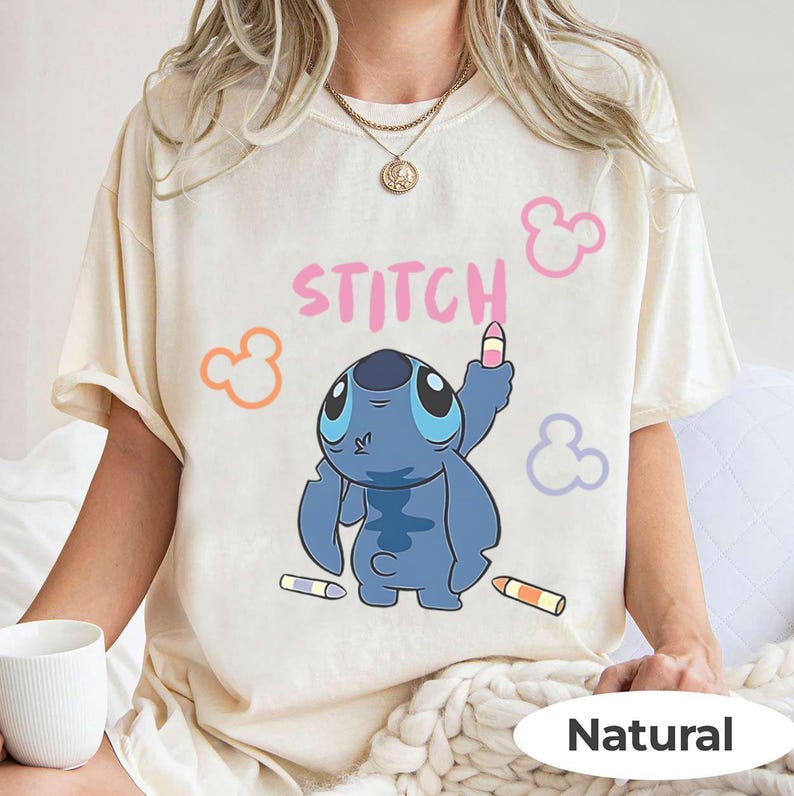 May include: Cream-colored t-shirt featuring a blue cartoon character holding a crayon, with the word "STITCH" in pink. The design includes colorful crayon accents and Mickey Mouse-like shapes. The shirt is labeled "Natural".
