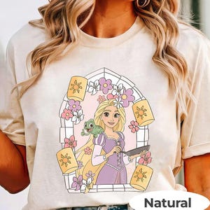 May include: Beige t-shirt featuring a cartoon illustration of a princess with long blonde hair, a green frog, and floating lanterns. The princess is wearing a purple dress and holding a pan. The design is framed by a floral archway. The word "Natural" is at the bottom.