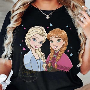 Puede incluir: Camiseta negra con una ilustraci&oacute;n de dibujos animados de dos personajes femeninos con cabello rubio y casta&ntilde;o rojizo, rodeados de estrellas rosas y azules. El texto "Duey World" y "Fashion and enjoyment" est&aacute; impreso debajo.