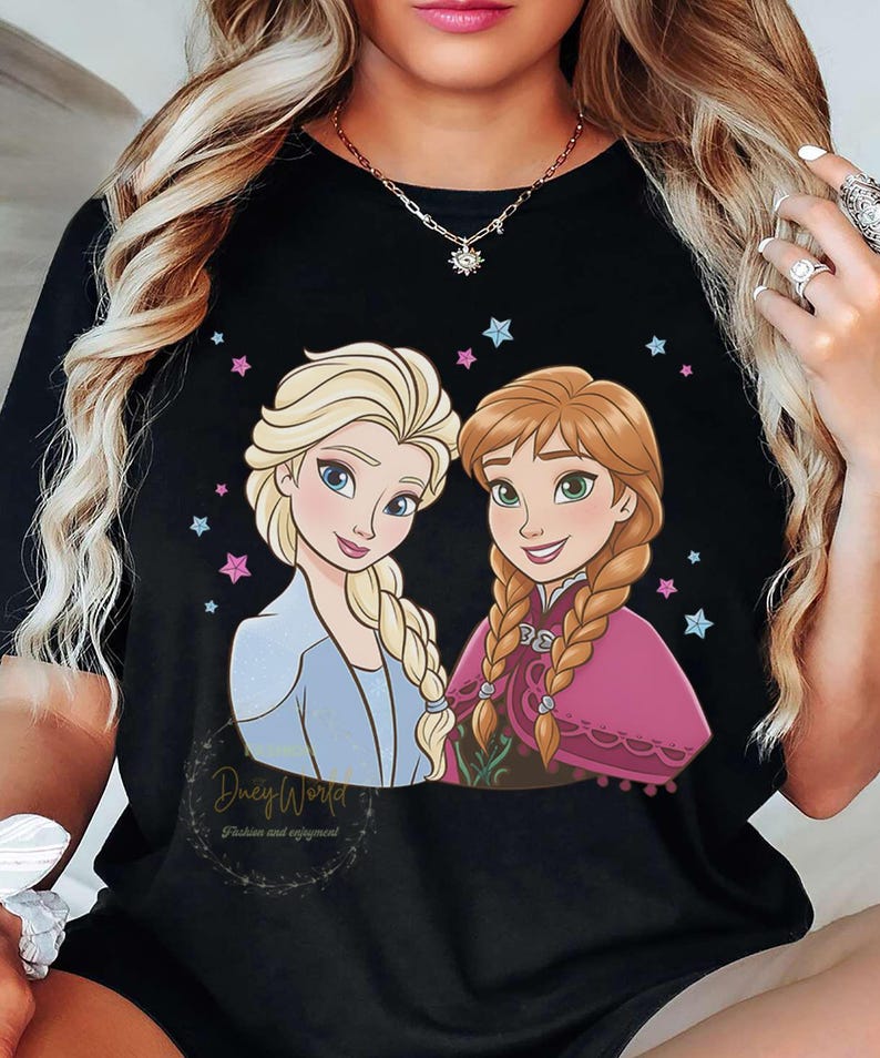 Puede incluir: Camiseta negra con una ilustraci&oacute;n de dibujos animados de dos personajes femeninos con cabello rubio y casta&ntilde;o rojizo, rodeados de estrellas rosas y azules. El texto "Duey World" y "Fashion and enjoyment" est&aacute; impreso debajo.