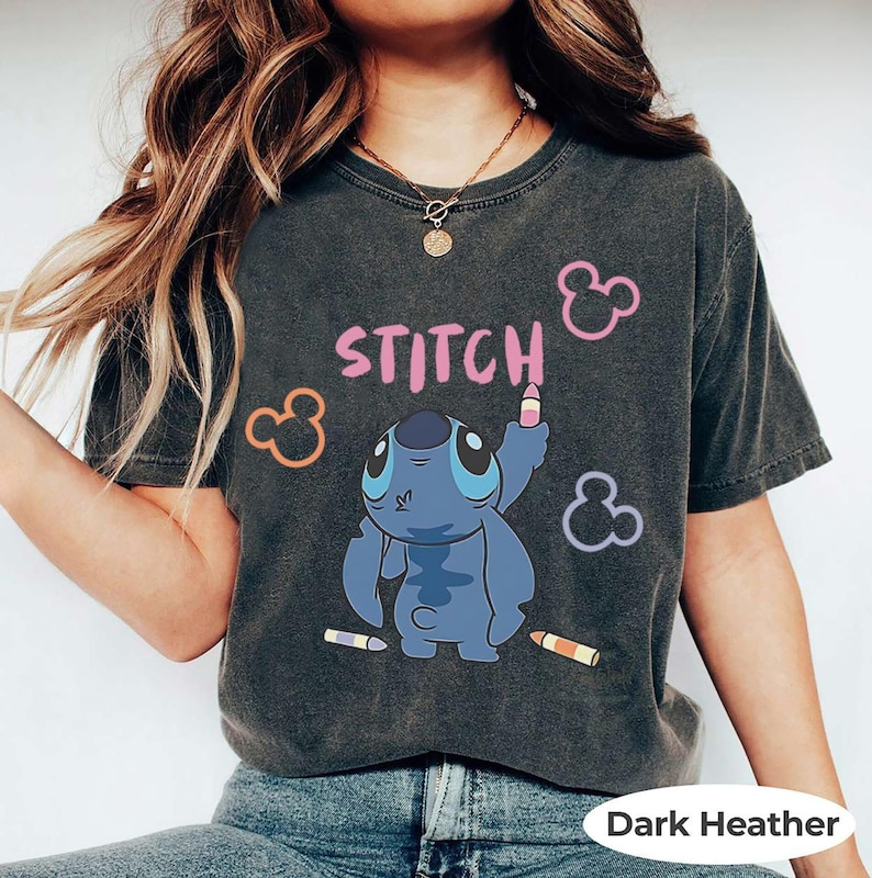 May include: Dark heather t-shirt featuring a cartoon Stitch character with the word "Stitch" in pink. Stitch is holding a crayon, with Mickey Mouse-shaped icons and crayons scattered around the design. The shirt is a dark gray color.