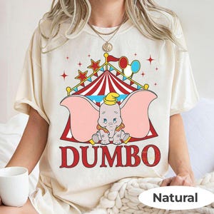 May include: Cream-coloured t-shirt featuring a graphic of Dumbo the elephant from the Disney film. The design includes a circus tent, balloons, and the word "DUMBO" in red. The shirt also has the word "Natural" printed on it.