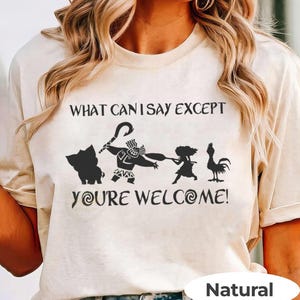 Puede incluir: Camiseta beige con la frase "WHAT CAN I SAY EXCEPT YOU'RE WELCOME!" y siluetas de personajes de la pel&iacute;cula Moana. La camiseta tiene cuello redondo y mangas cortas. La palabra "Natural" est&aacute; impresa en la parte inferior.