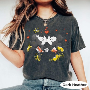 May include: Dark heather gray t-shirt featuring a graphic of Mickey and Minnie Mouse kissing, surrounded by red hearts and a yellow flower. The shirt has a vintage, worn look. The text "Dark Heather" is at the bottom.