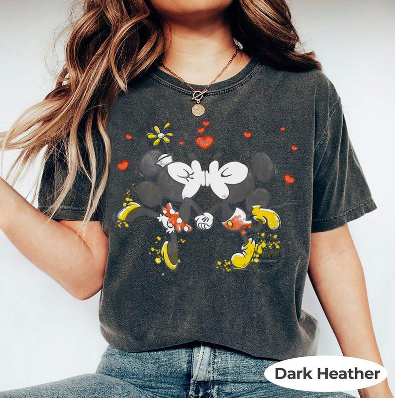 May include: Dark heather gray t-shirt featuring a graphic of Mickey and Minnie Mouse kissing, surrounded by red hearts and a yellow flower. The shirt has a vintage, worn look. The text "Dark Heather" is at the bottom.