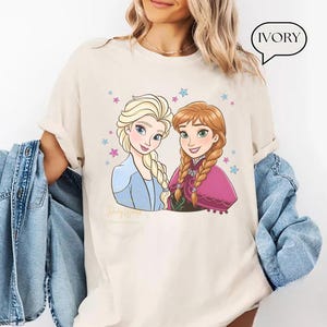 Puede incluir: Camiseta de color marfil con una ilustraci&oacute;n de dibujos animados de dos personajes femeninos con trenzas rubias y casta&ntilde;as, rodeadas de estrellas. Un personaje viste un atuendo azul, el otro una capa rosa. La palabra "IVORY" est&aacute; en una burbuja.