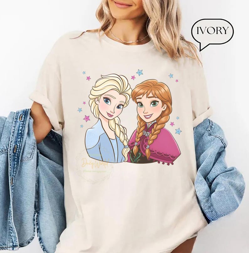Puede incluir: Camiseta de color marfil con una ilustraci&oacute;n de dibujos animados de dos personajes femeninos con trenzas rubias y casta&ntilde;as, rodeadas de estrellas. Un personaje viste un atuendo azul, el otro una capa rosa. La palabra "IVORY" est&aacute; en una burbuja.