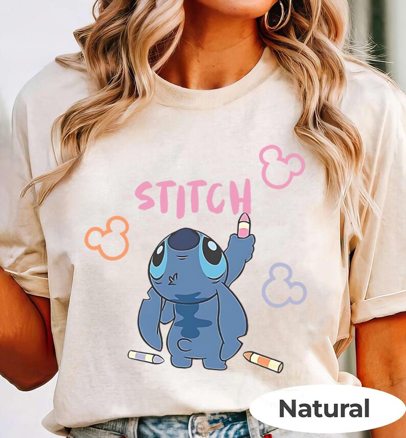 May include: Beige t-shirt featuring a blue cartoon character, with the word "STITCH" in pink above it. The character is holding a crayon, with additional crayons and Mickey Mouse-like shapes. The shirt has a "Natural" label.