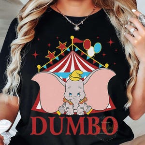 May include: Black t-shirt with a Dumbo graphic. The design features a circus tent, stars, and balloons. The word "DUMBO" is in red, with a cartoon elephant wearing a yellow hat. The shirt has a classic crew neck.