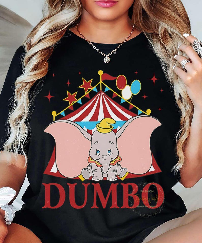 May include: Black t-shirt with a Dumbo graphic. The design features a circus tent, stars, and balloons. The word "DUMBO" is in red, with a cartoon elephant wearing a yellow hat. The shirt has a classic crew neck.