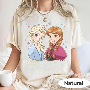 Puede incluir: Camiseta color crema con una ilustraci&oacute;n de dibujos animados de dos personajes femeninos con cabello rubio y casta&ntilde;o rojizo, rodeados de estrellas. La palabra "Natural" est&aacute; impresa en un &oacute;valo blanco en la parte inferior.