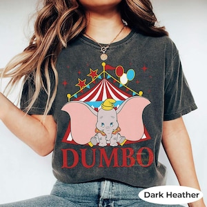 May include: Dark heather grey t-shirt with a Dumbo the elephant graphic in front of a circus tent. The word "DUMBO" is printed in red below the elephant. The shirt has a vintage, distressed appearance.