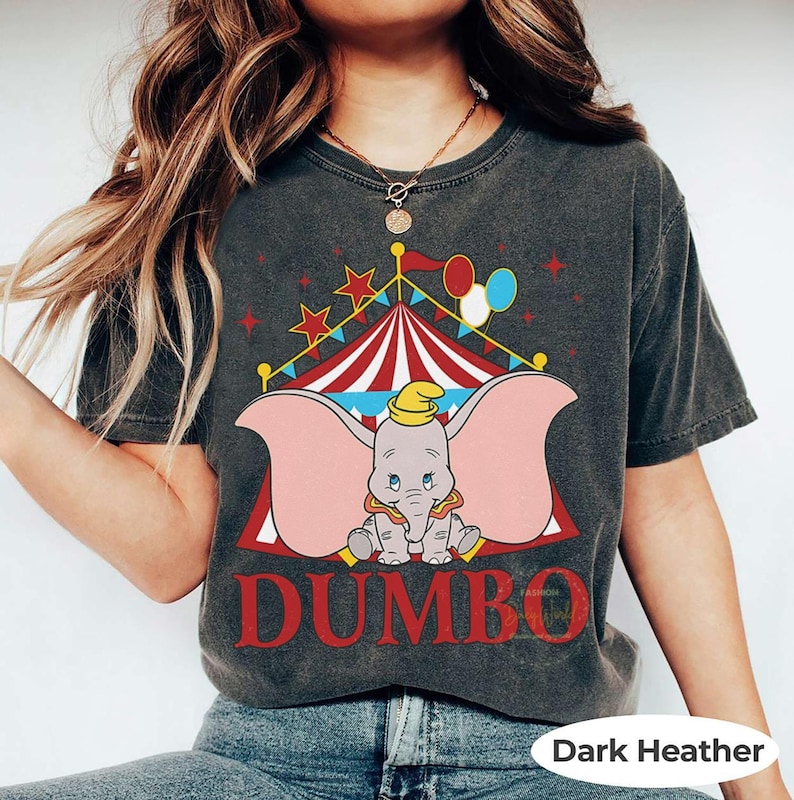 May include: Dark heather grey t-shirt with a Dumbo the elephant graphic in front of a circus tent. The word "DUMBO" is printed in red below the elephant. The shirt has a vintage, distressed appearance.
