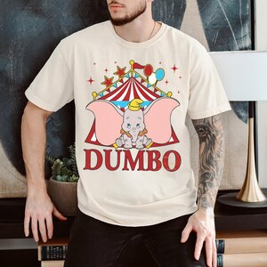 May include: Cream-coloured t-shirt featuring a Dumbo graphic. The design includes a circus tent, stars, balloons, and the word "DUMBO" in red. The cartoon elephant is wearing a yellow hat and has large pink ears.