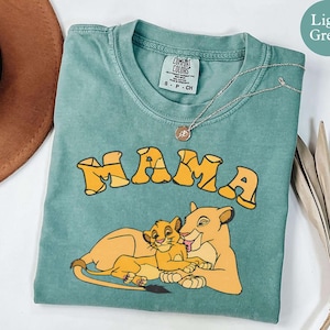 May include: Light green t-shirt with the word "MAMA" in yellow, along with a cartoon illustration of a lioness and cub. The shirt has a crew neck and a small tag. A silver necklace with a pendant is also visible.