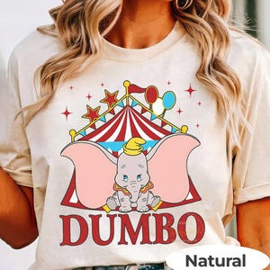 May include: Beige t-shirt with a Dumbo graphic. The design features a circus tent, stars, and balloons. The word "DUMBO" is printed in red. The shirt is made of a soft, natural-coloured fabric.