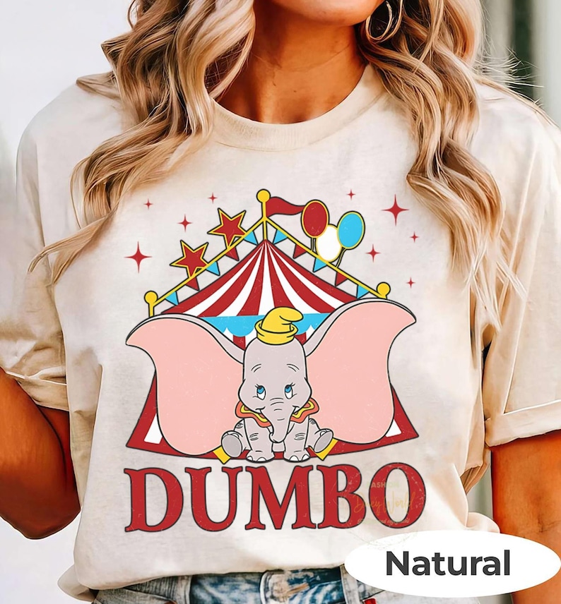 May include: Beige t-shirt with a Dumbo graphic. The design features a circus tent, stars, and balloons. The word "DUMBO" is printed in red. The shirt is made of a soft, natural-coloured fabric.