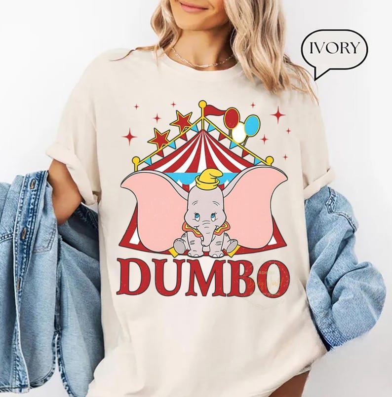 May include: Ivory t-shirt with a Dumbo graphic. The design features a circus tent, stars, and balloons. The word "DUMBO" is printed in red, with a cartoon elephant in the center. This casual t-shirt is suitable for everyday wear.