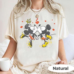 May include: Cream-colored t-shirt featuring a cartoon illustration of Mickey and Minnie Mouse kissing, with red hearts and a flower. The word "Natural" is printed at the bottom. The shirt has short sleeves.