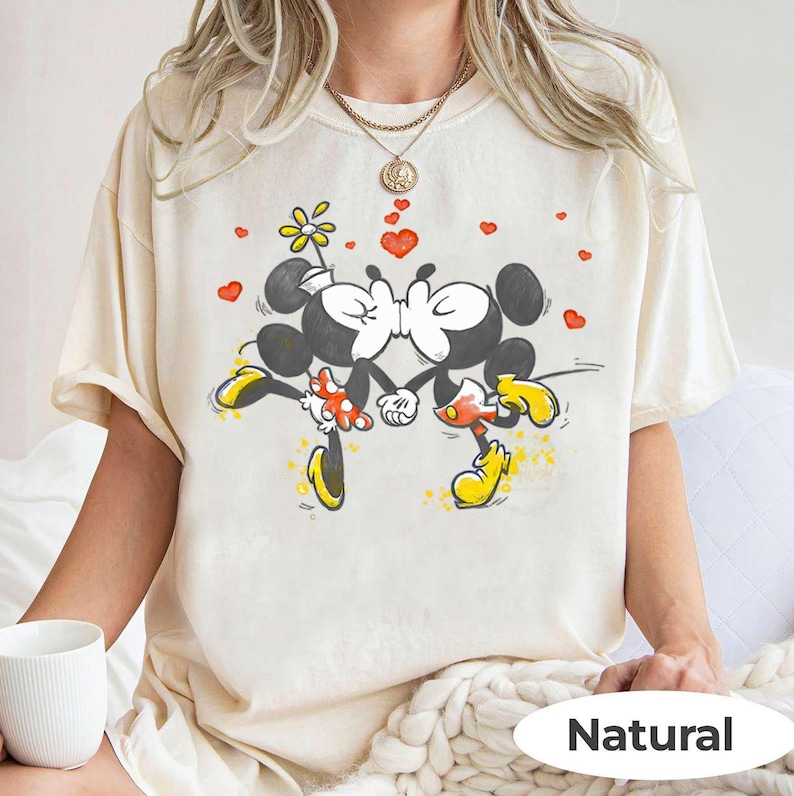 May include: Cream-colored t-shirt featuring a cartoon illustration of Mickey and Minnie Mouse kissing, with red hearts and a flower. The word "Natural" is printed at the bottom. The shirt has short sleeves.