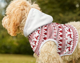 Puede incluir: Un perro con un suéter con capucha blanco con un patrón navideño rojo y blanco. El patrón incluye renos, árboles y copos de nieve. El perro está sobre hierba verde.