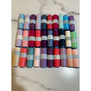 May include: A collection of thirty-three cylindrical spools of polyester yarn arranged in a grid. Each spool features a white paper label with the text Polyester Yarn Samoilov Inc. and a specific color number, showcasing a wide variety of colors.