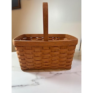 May include: A rectangular woven basket with a tall, arched handle. The basket is made of light brown wood and has a square base. The handle is attached to the top of the basket. The basket is empty.