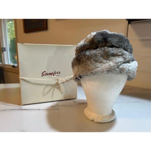 May include: A gray and white faux fur hat with a textured, multi-tonal appearance sits on a white mannequin head. Beside it is a square, off-white cardboard box tied with a thin string, featuring the red text Stampfer's printed on the front.