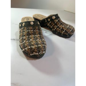 May include: A pair of slip-on clogs featuring a multi-colored tweed fabric upper in shades of olive green, black, and tan. The dark green trim is embroidered with small floral patterns and metallic studs secure the sole to the upper.