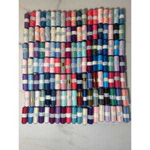 Op de afbeelding: Een grote collectie naaigarenklosjes gerangschikt in een rasterpatroon. Elke klos heeft een eigen effen kleur, waaronder tinten blauw, roze, paars, groen, rood en neutrale tinten, met witte labels waarop de kleurnummers staan vermeld.