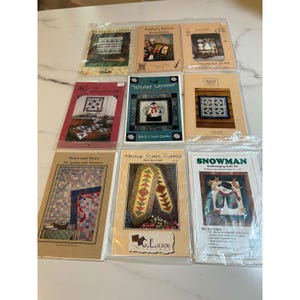 May include: A collection of quilt kits in plastic packaging. The kits feature various designs, including a snowman, floral patterns, and geometric shapes. Some kits have text such as "Winter Layover" and "Snowman".