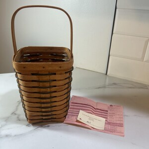 May include: A rectangular woven basket with a handle, in light brown with dark brown trim. The basket has a curved handle. A pink paper with text is next to the basket.