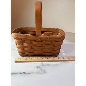 May include: A rectangular, woven wooden basket with a tall, arched handle. The basket is light brown and has a ruler next to it. The basket is approximately 10 inches long.